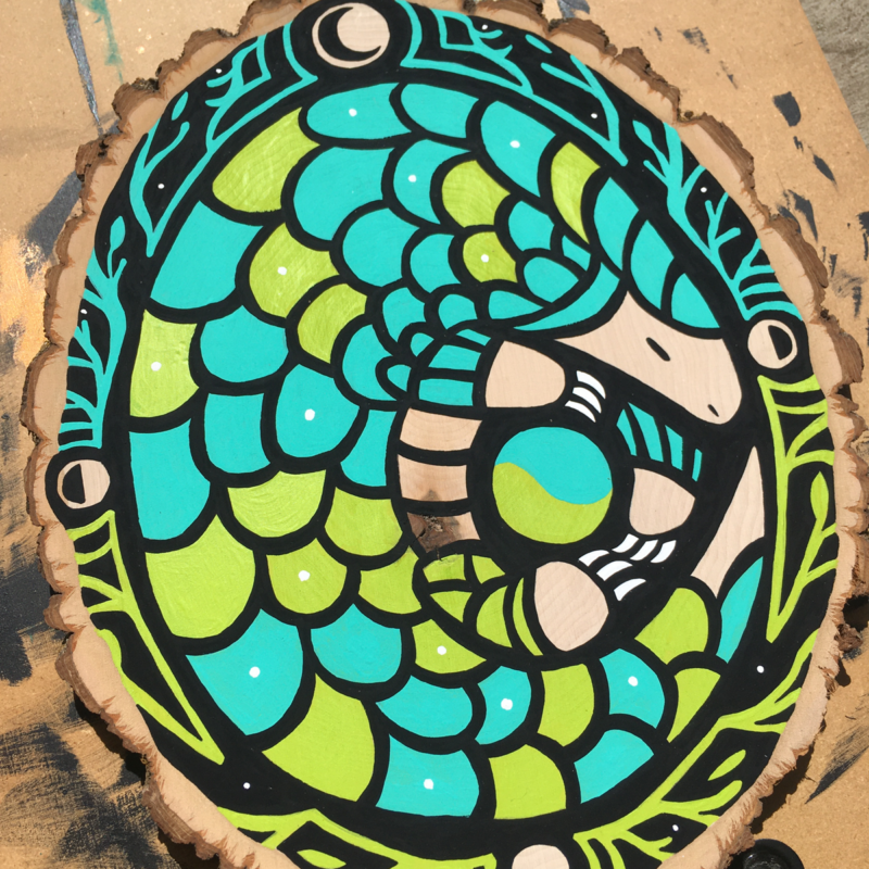 A vibrant pangolin painted with a bright turquoise and lime green sits curled up in the center of a decorative border consisting of vines, leaves, and phases of the moon on a black background.