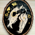 A painting on a wood slice featuring a bear skull against a black background with golden poppies entangled around the skull.