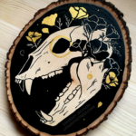 A painting on a wood slice featuring a bear skull against a black background with golden poppies entangled around the skull.