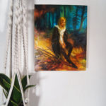 Painting of a girl Wearing a cloak sitting in a golden landscape ahead of a manganese forest. The Painting is hung next to a plant in a macrame hanger.