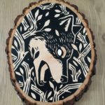 A painting on a wood slice featuring a wolf encircled within branches. It is diving down, in the center of the piece, with it's mouth open.