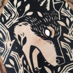 A painting on a wood slice featuring a wolf encircled within branches. It is diving down, in the center of the piece, with it's mouth open.