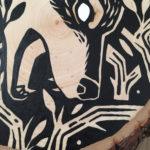 A painting on a wood slice featuring a wolf encircled within branches. It is diving down, in the center of the piece, with it's mouth open.