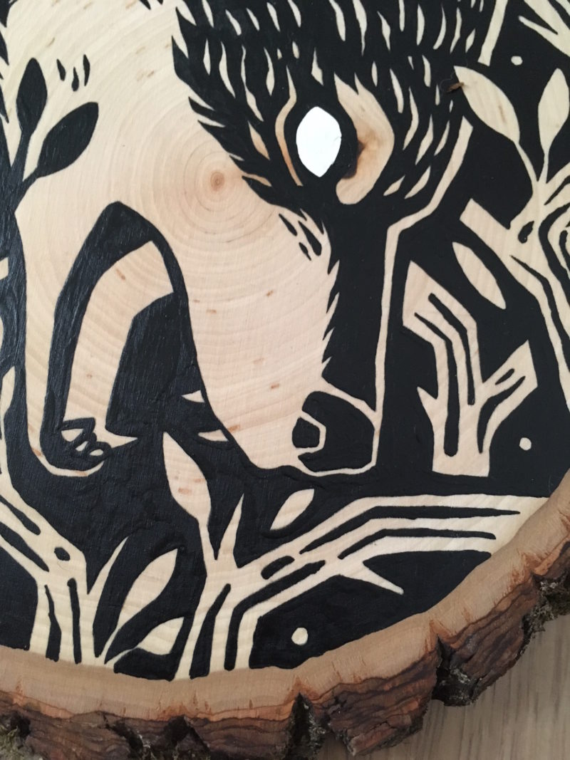 A painting on a wood slice featuring a wolf encircled within branches. It is diving down, in the center of the piece, with it's mouth open.