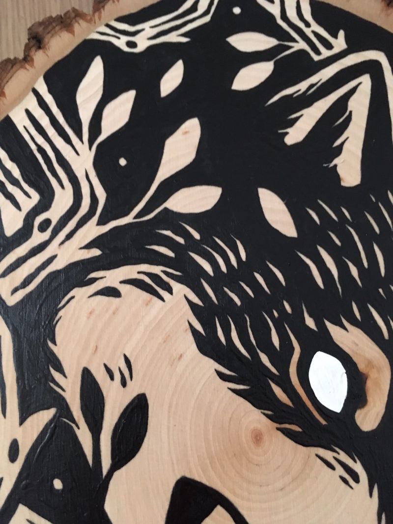 A painting on a wood slice featuring a wolf encircled within branches. It is diving down, in the center of the piece, with it's mouth open.
