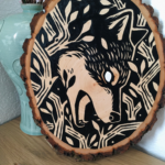 A painting on a wood slice featuring a wolf encircled within branches. It is diving down, in the center of the piece, with it's mouth open.