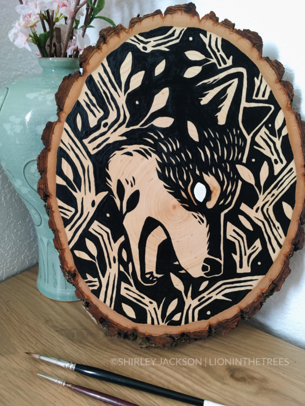 A painting on a wood slice featuring a wolf encircled within branches. It is diving down, in the center of the piece, with it's mouth open.