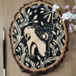 A painting on a wood slice featuring a wolf encircled within branches. It is diving down, in the center of the piece, with it's mouth open.
