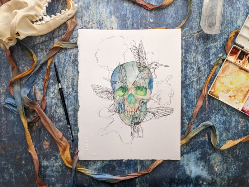 This is an image of a blue watercolor skull painting that has glowing green eyes. The skull is a darker blue around the edges and gradually gets lighter and warmer around the eye sockets and nasal cavity, with a rusty orange mixing in the cooler colors. The is an overlay of three ink hummingbirds drawn in a scribbly fashion directly over the skull. The hummingbirds are connected with tangled ink linework that guides the eye around the piece.