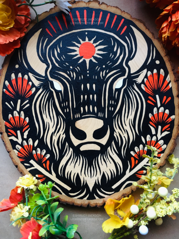 A painting on a wood slice featuring an American Bison with a red sun symbol on its forehead. It is also surrounded by Indian Paintbrush aka Prairie-Fire plants on either side.