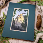 Golden Hour dragon standing on a cliff with wings out above a forest painted in watercolor photo of artwork in mat at an angle