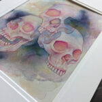 Pictured is a watercolor painting of three overlapping skulls floating amidst rich watercolor washes.