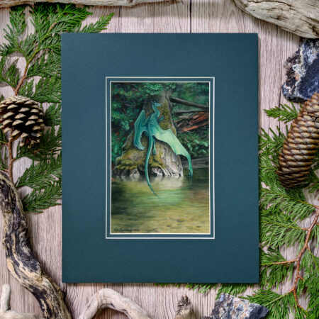 A dragon lays on a shaded mossy log above a pond. Photo of the artwork in the mat on a table surrounded by pine cones and cedar branches