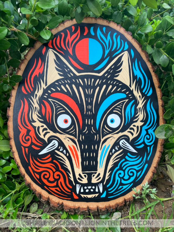 A painting on a wood slice featuring a coyote head. The left side is coloured with red details, and the right side is coloured with light blue details.