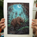 photograph of hands holding an ink and watercolor illustration of a brown bear sleeping peacefully inside a cave with a crescent moon and stars overhead.