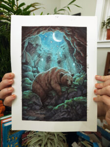 photograph of hands holding an ink and watercolor illustration of a brown bear sleeping peacefully inside a cave with a crescent moon and stars overhead.