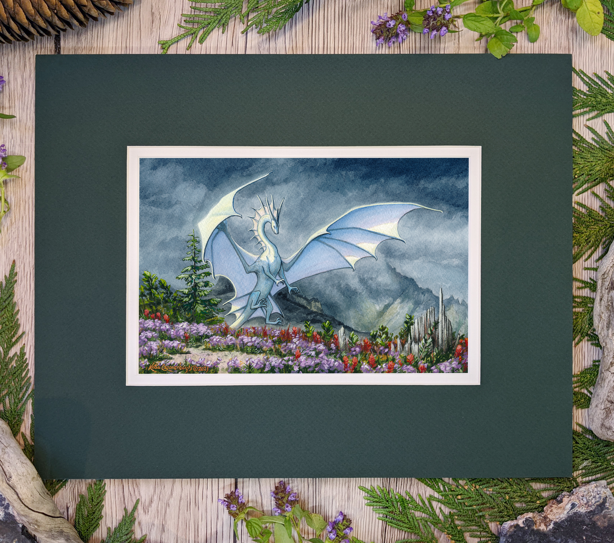 Photo of the artwork with mat. The bottom mat is white, the top main mat is a deep muted forest green. There are plants and leaves laid around the artwork on a table. The artwork is a white dragon alighting on a hilltop covered in purple and red flowers. The sky is dark and rainy, some sunlight reaches the mountain in the background. The dragon is white, the sun pierces the clouds to light it as it spreads its wings to land in the flowers.