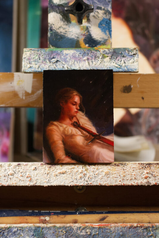 A miniature oil painting of a girl lying down, looking at the sword resting over her chest and stomach.