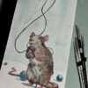 Painting of playful rat holding blue string with teal beads around the floor. Artist paintbrushes to the right of the work.