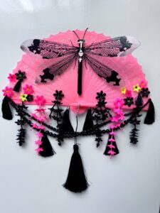 A shiny black dragonfly rests on a bright pink vintage fan decorated with a lot of fringe, beads and cascading flowers