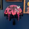 A shiny black dragonfly rests on a bright pink vintage fan decorated with a lot of fringe, beads and cascading flowers against a blue wall