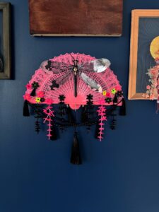 A shiny black dragonfly rests on a bright pink vintage fan decorated with a lot of fringe, beads and cascading flowers against a blue wall