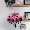 A shiny black dragonfly rests on a bright pink vintage fan decorated with a lot of fringe, beads and cascading flowers sits on a shelf with other art around it