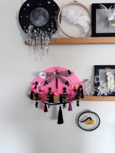 A shiny black dragonfly rests on a bright pink vintage fan decorated with a lot of fringe, beads and cascading flowers sits on a shelf with other art around it