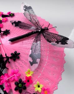 Detail of shiny black dragonfly rests on a bright pink vintage fan decorated with a lot of fringe, beads and cascading flowers