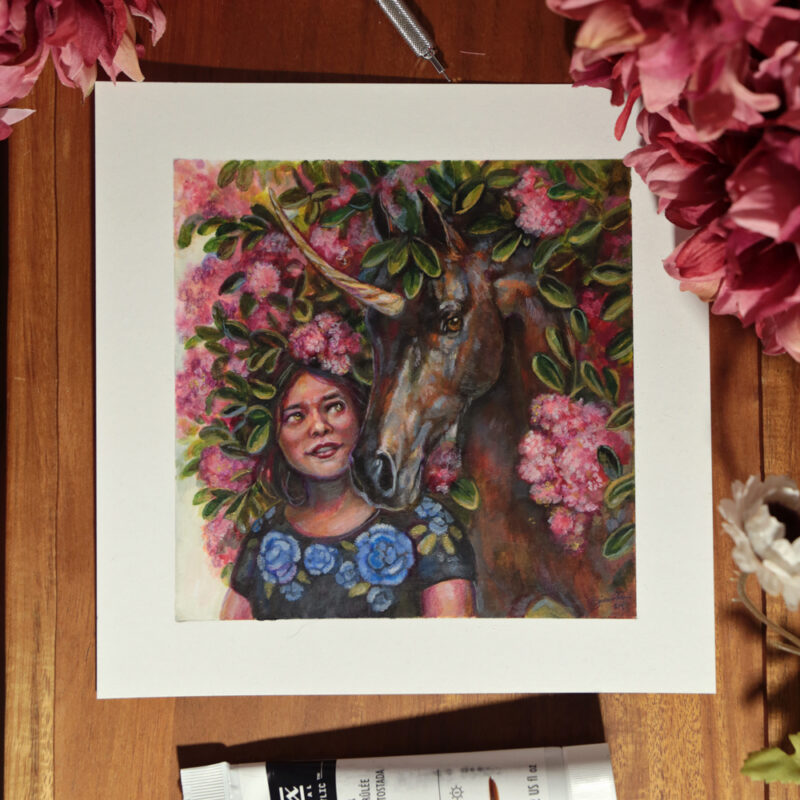 A mixed media painting of a woman or female representing character next to an unicorn. The unicorn is dark brown with a hand embellished gold paint horn. They're surrounded by bunches of pink flowers and green leaves. This is a photograph from above. Taken at night time