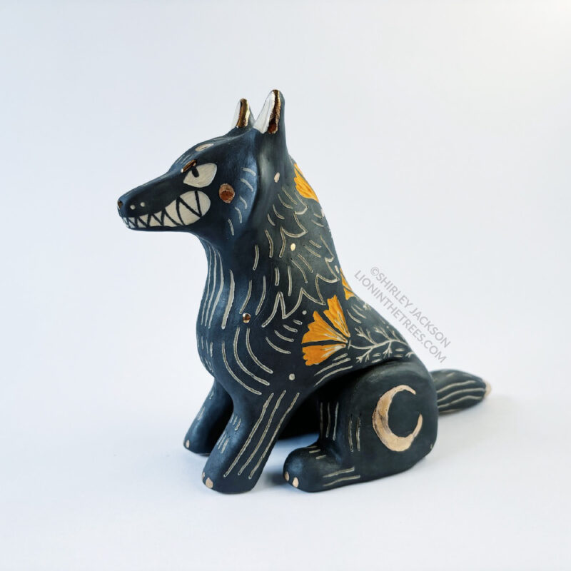 Sgraffito wolf totem done with black and orange underglazes, and finished with gold overglaze in some areas. The wolf is sitting down and California Poppies have been carved into it's side along with other markings. There are also crescent moon details done in gold overglaze. This photo shows the 3/4 left front view.