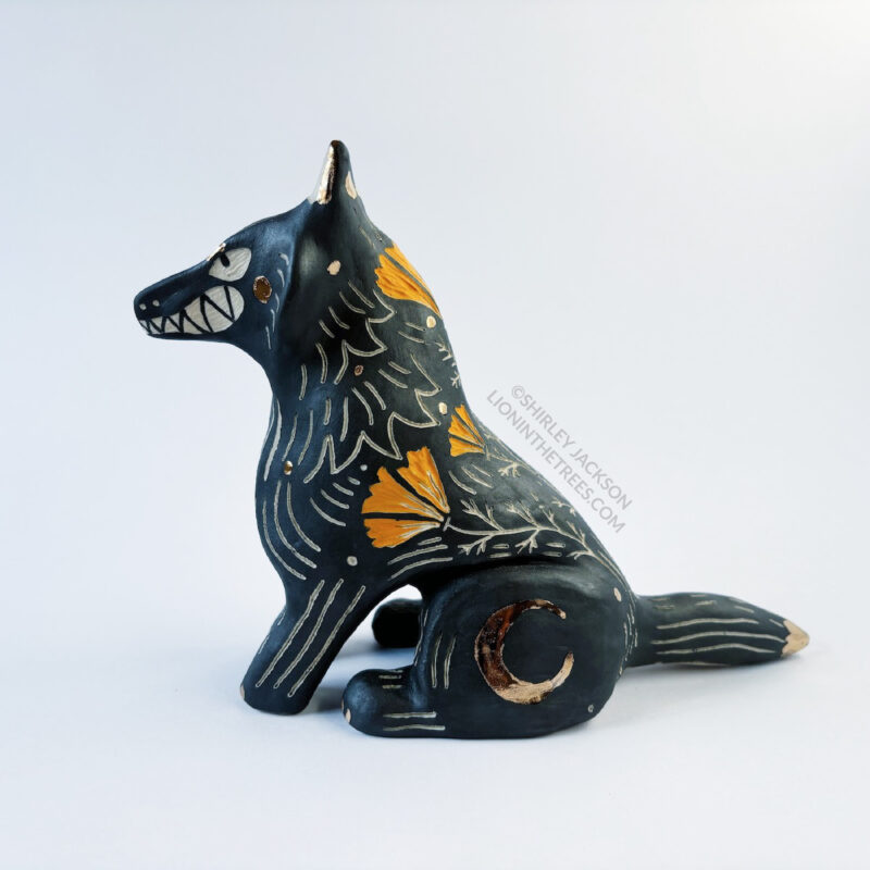 Sgraffito wolf totem done with black and orange underglazes, and finished with gold overglaze in some areas. The wolf is sitting down and California Poppies have been carved into it's side along with other markings. There are also crescent moon details done in gold overglaze. This photo shows the left side of the sculpture.