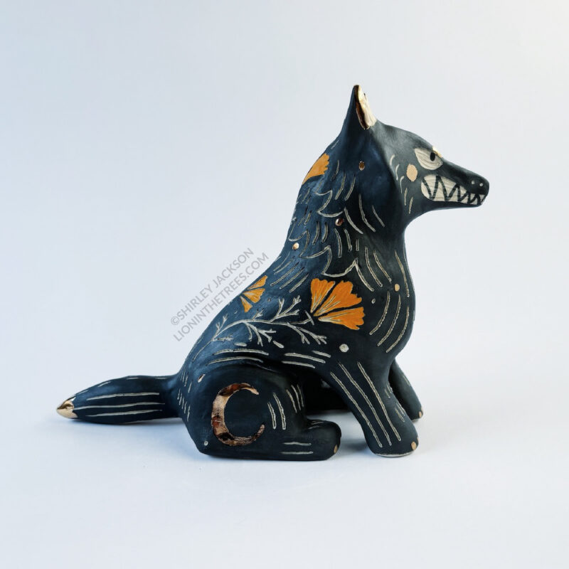 Sgraffito wolf totem done with black and orange underglazes, and finished with gold overglaze in some areas. The wolf is sitting down and California Poppies have been carved into it's side along with other markings. There are also crescent moon details done in gold overglaze. This photo shows the right side of the sculpture.