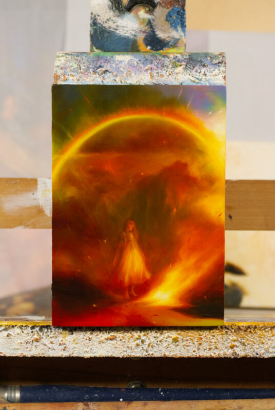 A photo from afar of an oil painting containing a young girl standing by a raging fire, and sparkling arch of a rainbow overhead