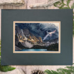 Flat scan view of a white dragon flying over a lake similar to Moraine Lake in Banff, Canada. A dark storm rises from behind the mountains, lit in a blazed line across the center from the setting sun. Image shows the artwork in the mat, a tawny brown inner mat and a forest green top mat.