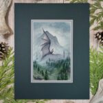 A dark gray dragon rises up through the foggy forested hillside with a mountain and cloudy sky in the background. Photo shows artwork in the mat it will come in.