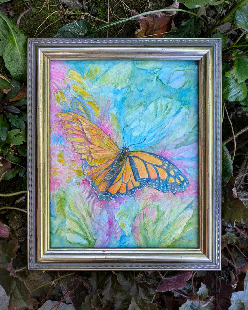 Photo of a butterfly painting. It is framed in pale gold and surrounded by leaves outside. The butterfly is a bright orange monarch.