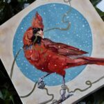Original oil painting by Catherine Moore depicting a red cardinal holding a gold string against a circular shape of blue with snow falling
