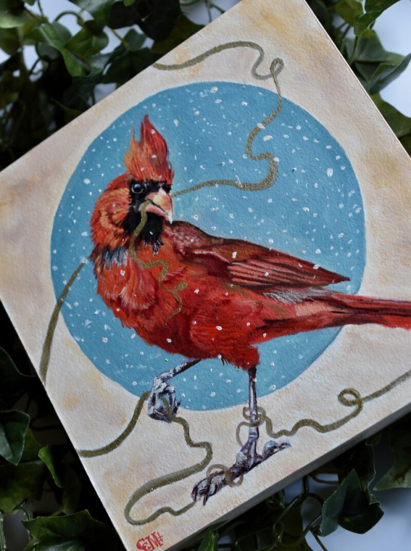 Original oil painting by Catherine Moore depicting a red cardinal holding a gold string against a circular shape of blue with snow falling