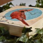 Original oil painting by Catherine Moore depicting a red cardinal holding a gold string against a circular shape of blue with snow falling