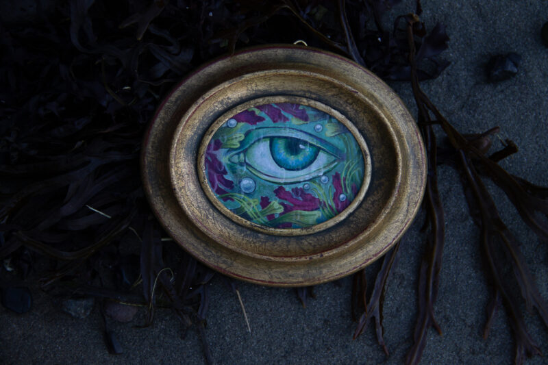 A small watercolour and gouache painting of an underwater eye in a gold frame. Nestled on a bed of seaweed.