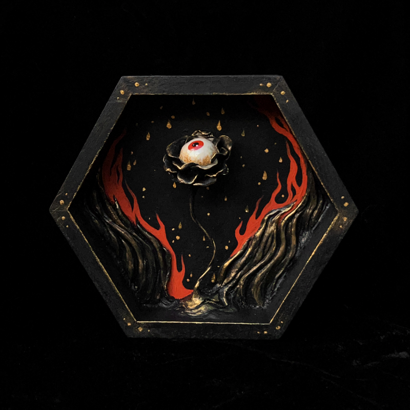 A sculpture of a flower with an eyeball surrounded by flames contained in a hexagonal wooden shadowbox.