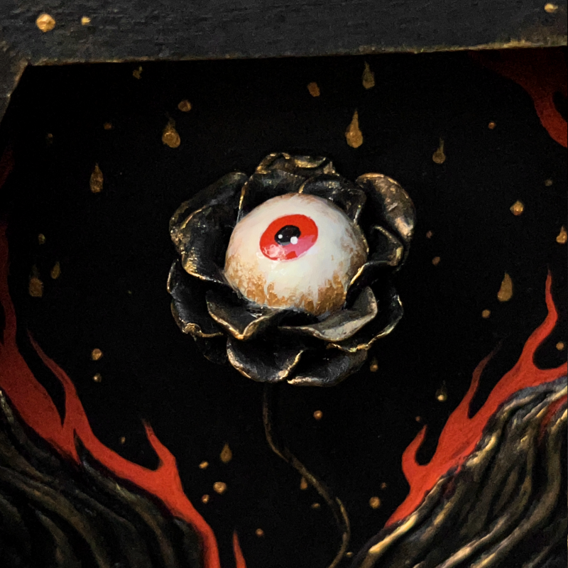 A sculpture of a flower with an eyeball surrounded by flames contained in a hexagonal wooden shadowbox.