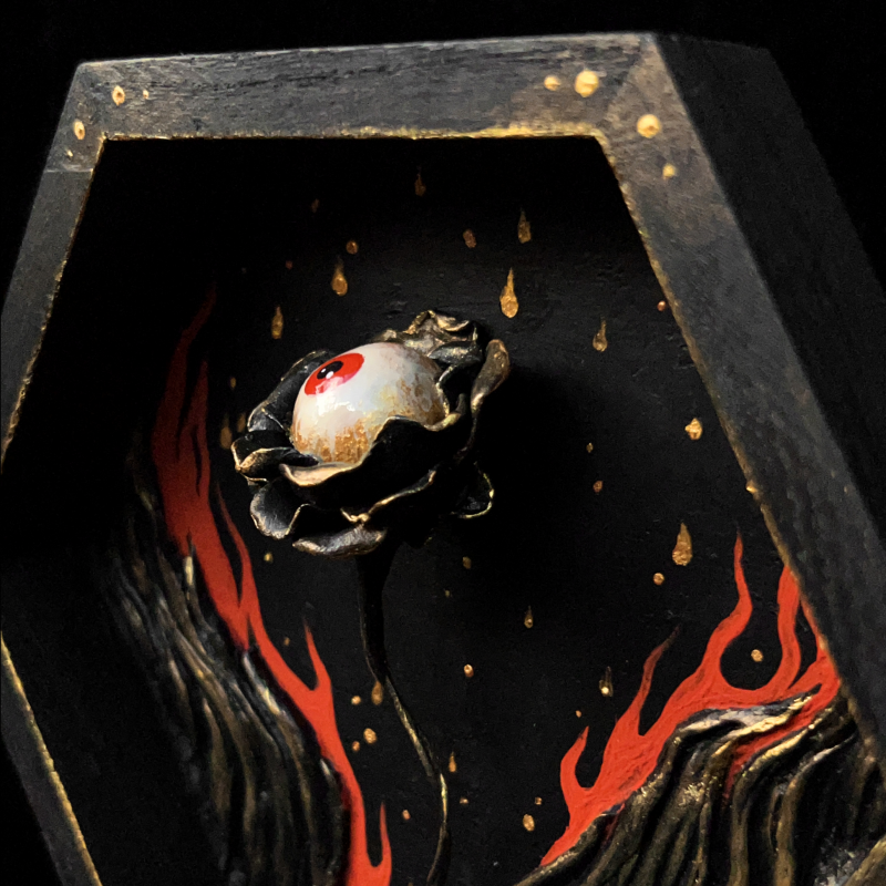 A sculpture of a flower with an eyeball surrounded by flames contained in a hexagonal wooden shadowbox.