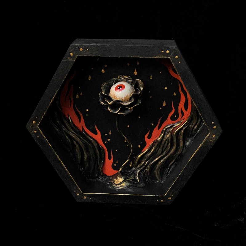 A sculpture of a flower with an eyeball surrounded by flames contained in a hexagonal wooden shadowbox.