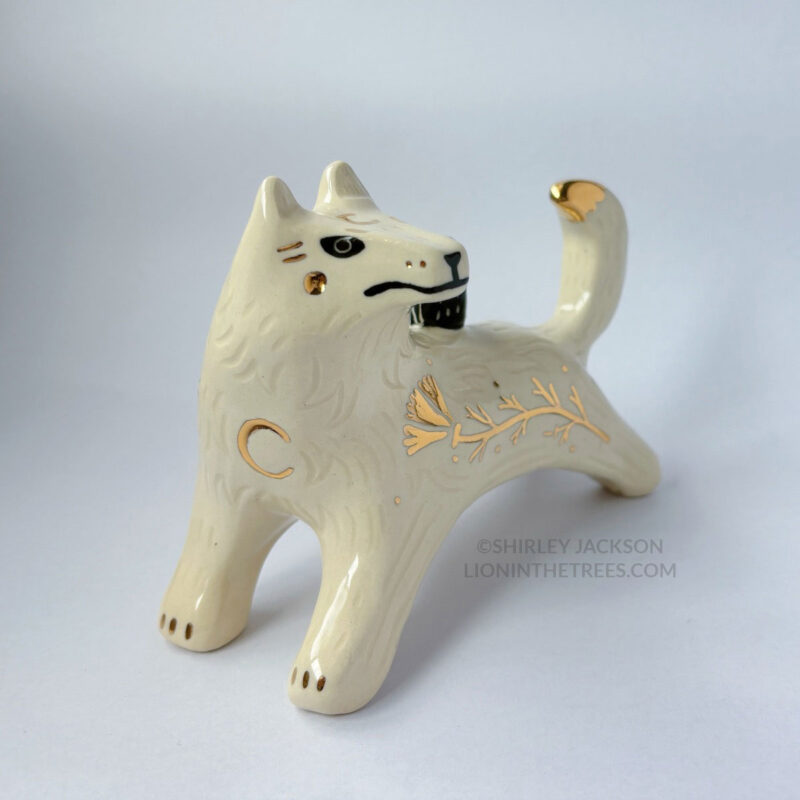 White wolf totem in an outstretched, running position done with minimal black underglaze, finished with clear glaze and gold overglaze details such as a crescent moon on it's chest, forehead, and gold California Poppy motifs on it's side.