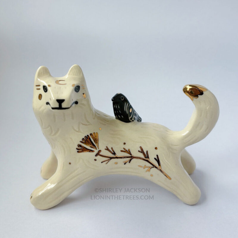 White wolf totem in an outstretched, running position done with minimal black underglaze, finished with clear glaze and gold overglaze details such as a crescent moon on it's chest, forehead, and gold California Poppy motifs on it's side. There is also a small, black raven sitting on their back.