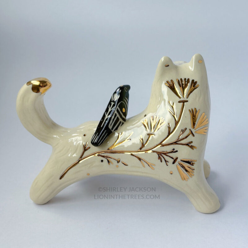 White wolf totem in an outstretched, running position done with minimal black underglaze, finished with clear glaze and gold overglaze details such as a crescent moon on it's chest, forehead, and gold California Poppy motifs on it's side. There is also a small, black raven sitting on their back. The back view of the sculpture shows a large California Poppy carved into it's side done with gold luster.