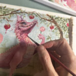 Picture of artist working on a painting of a pink dragon. The artist's hand is shown holding a paint brush.