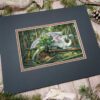 The matted painting sitting on a table with pine cones and wood sticks. A white dragon laying on fallen trees in the forest.
